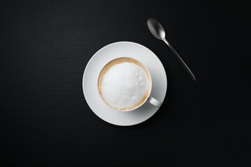 White cup of cappuccino on a black textured background. Coffee on black. Close-up on a white cup. Cappuccino Flat lay, black background, white cup with spoon.