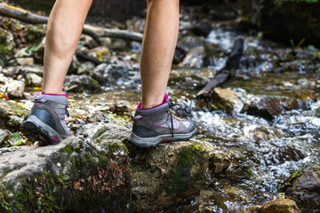 Woman with hiking boots walking on rocks in river. Tourist trekking at mountain trail