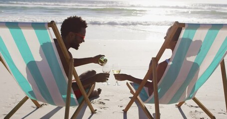 African american couple drinking wine together sitting on deck chairs at the beach - Powered by Adobe