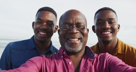 Smiling african american senior father and twin teenage sons standing on a beach taking a selfie - Powered by Adobe
