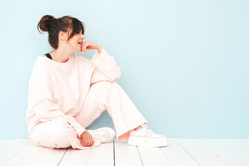 Portrait of young beautiful smiling female in trendy summer pink clothes. Sexy carefree woman sitting near light blue wall in studio. Positive model having fun indoors. Cheerful and happy