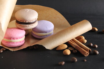 Three multi-colored macaroons lie on crafted brown paper next to lie almond sticks cinnamon and coffee on a black background with place for text and with a copyspace
