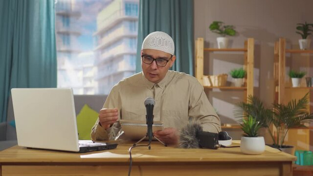 Muslim man in a skullcap records a podcast while sitting in front of a microphone in living room