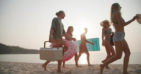 Dolly shot of young caucasian men and women walk on the beach in slow motion. Group of friends carry a cool box and swim ring with happy emotion in summertime. Travel, vacation and friendship concept. - Powered by Adobe