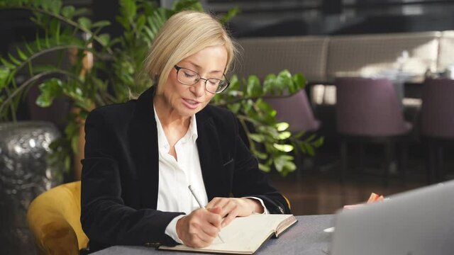 Business lady sitting at a table during business meeting in cafe, restaurant, holding eyeglasses in hands, taking notes in notebook, smiling.