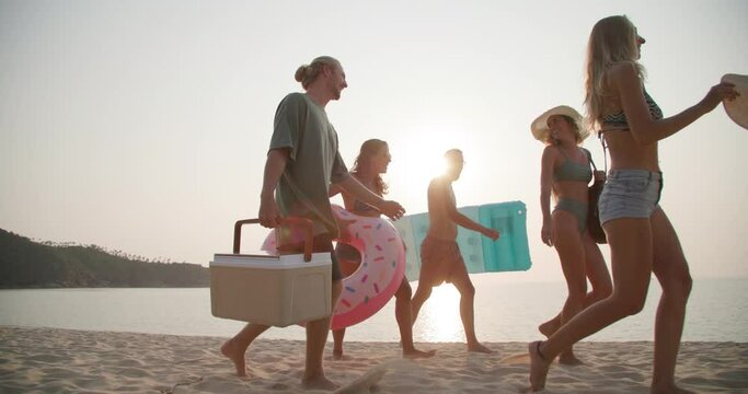 Dolly shot of young caucasian men and women walk on the beach in slow motion. Group of friends carry a cool box and swim ring with happy emotion in summertime. Travel, vacation and friendship concept.