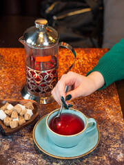 Fruit red tea is poured into a mug. A woman's hand is stirring sugar with a spoon. Next to it is a chaikik fren press and a plate of white and cane sugar. Vitamin teas.