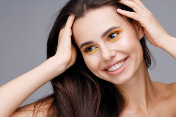 Portrait of a charming girl with yellow bright makeup, lush eyebrows and plump lips touches her hair. Personal care, cosmetology and makeup concept. Woman expresses joyful emotions.
