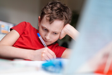 Naklejka na ściany i meble Attentive focused hardworking schoolboy boy sits down at the table and does homework, looks at the textbook, at home. Back to school. The child does math, thinks about the solution of the problem