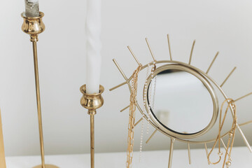 Modern gold boho mirror with gold necklace and vintage candles on white table. Jewellery and details