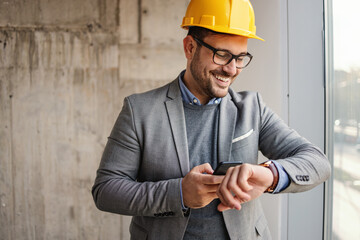 Smiling businessman with helmet standing in building in construction process next to a window, looking at wristwatch and holding phone. All works have been done on time.