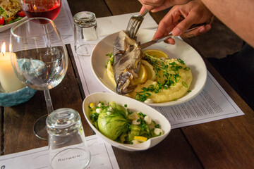 Restaurant setting. Man eating mashed potatoes and fried fish with fresh cucumber salad. Wine glass and white plates on a food menu.