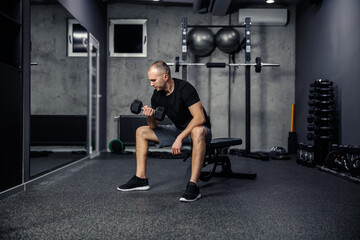Fototapeta na wymiar Exercises for shoulders and arms. Shot of the back of a middle-aged man holding and lifting a dumbbell with one hand while sitting on a sports bench indoor modern gym. Physical exercise, fitness