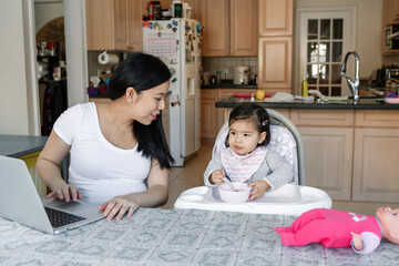 Asian Chinese young mother with daughter baby working online from home on Internet. Workplace of freelancer woman with kid girl. Stay home single mom working distant job. New normal. - 426180224