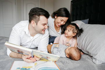 Asian Chinese mom, Caucasian father lying on bed with toddler girl and newborn baby. Multiracial family reading book, having fun, spending time together at home. Family authentic candid lifestyle.