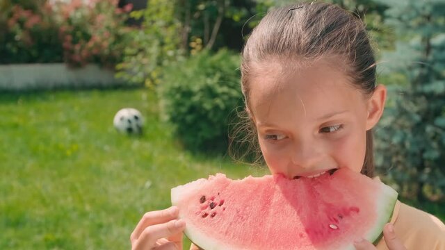Closeup portrait of cute 10-year-old girl eating juicy watermelon and smiling to camera standing in backyard on sunny summer day