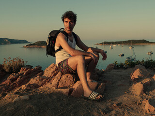 young man sitting on a stone above a beautiful  sardinian beach with boats and yachts
