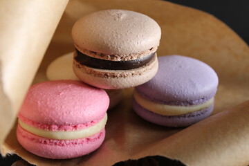 Three multi-colored macaroons lie on crafted brown paper next to lie almond sticks cinnamon and coffee on a black background with place for text and with a copyspace