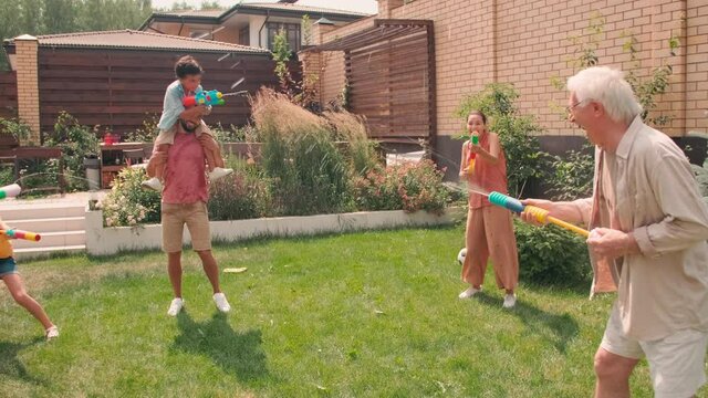 Sequence of footages of big multigenerational family with two elementary age kids spending summer day together in backyard shooting water guns splashing water on each other having fun