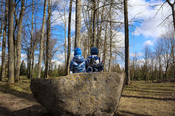Obraz premium Two children in blue jackets sit on a large boulder in an old park on a spring day, side view