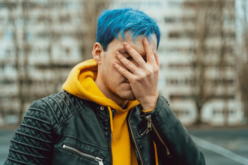 Portrait of young man closing his face with hand on background of high-rise building. Handsome guy with blue hair posing on city street in springtime.