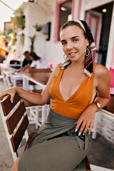 Charming tanned woman with wonderful smile, stylish scarf on the head waiting for breakfast widely and poses outside in street cafe.