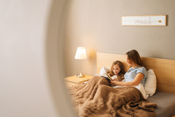 Reflection in mirror of young happy mother and adorable cheerful daughter reading children book...