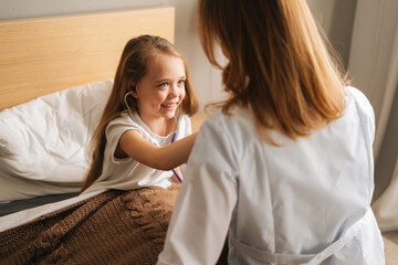 Close-up of happy little girl listening heartbeat with stethoscope to female doctor sitting in bed. Cute child playing as doctor nurse listening physician with stethoscope. Concept of children health