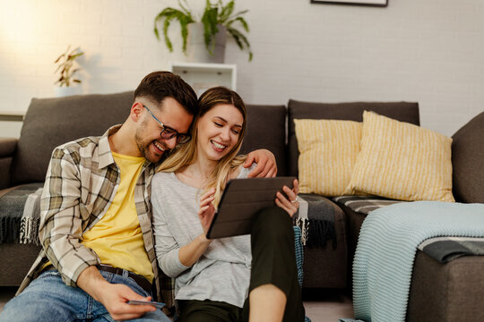 Happy young couple sitting on the floor and doing online shopping at home, using tablet computer.