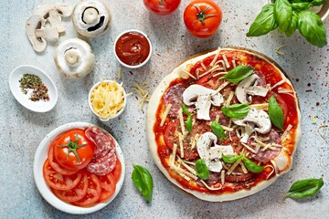 Cooking Italian pizza with tomato sauce, fresh tomatoes, cheese, mushrooms, salami slices  and basil . Fresh food cooking concept at home. Semi-finished product, raw dough.