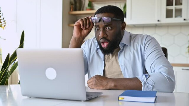 Shocked amazed African American bearded businessman or freelancer, sits at his desk, looks in surprise at his laptop, reads unexpected news, take off glasses, confused face expression