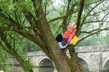 Muslim girl smiling and holding the book while sitting on tree branch