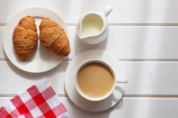 Cup of coffee with milk and two croissants on a plate on white wooden table with light shadow. Cozy morning flat lay. Top view