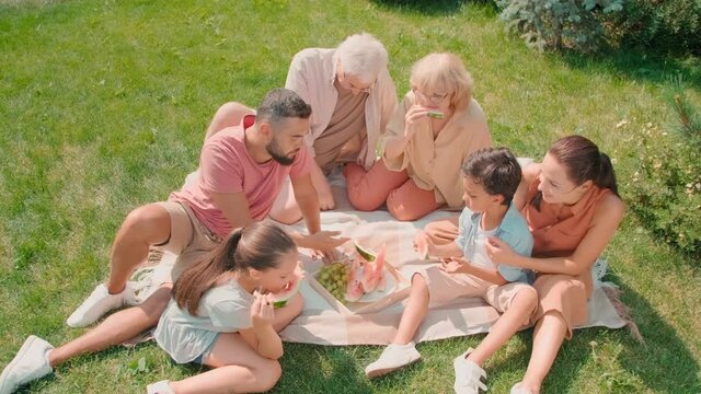 High-angle Shot Of Relaxed Multigenerational Family Of Six With Two Elementary Age Kids Spending Summer Day Together Sitting In Backyard Chatting And Eating Watermelon Enjoying Sunshine