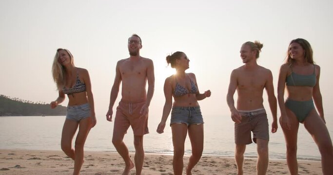 Dolly shot of young caucasian men and women walk and talk on the beach in slow motion. Group of friends in swimwear with happy emotion in summertime. Travel, vacation and friendship concept.