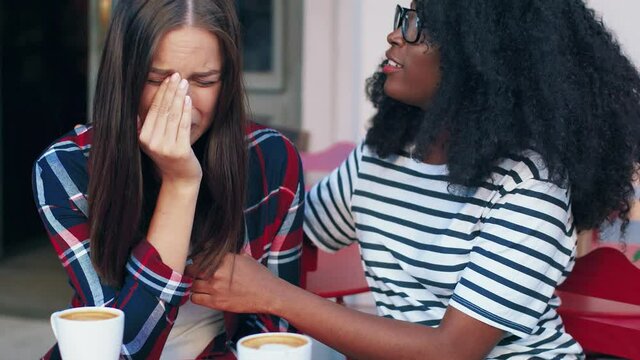 Two girlfriends are sitting on terrace of cafe and talking. Young african american girl calm friend stroking hand and hugging.