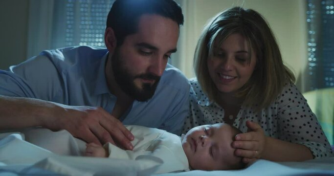 Cinematic shot of happy family, neo parents caressing and kissing with affection their newborn baby boy while sleeping peacefully with sweet dreams in a crib with soft blanket in a nursery at night. 