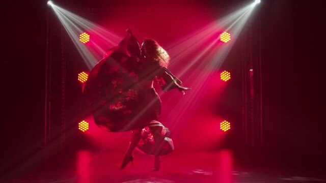 Silhouette of a passionate young woman in a gypsy dress whirls dancing and waving a long skirt. Female dancer dancing romany folk dance in dark smoky studio with red light. Slow motion.