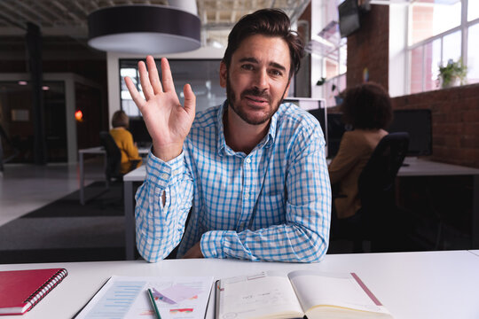 Caucasian businessman sitting at desk with documents saying hello having video call