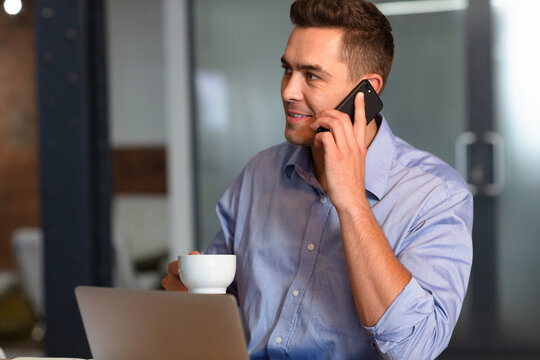 Happy casual caucasian businessman talking on smartphone drinking coffee sitting at desk