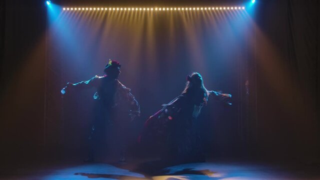 Silhouette couple dancers in romany clothes dancing in dark studio against background of bright lights. Man and woman passionately dancing gypsy dance performing in theatrical show. Slow motion.