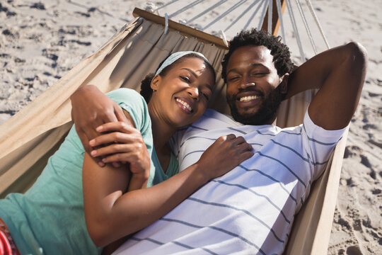 Happy african american couple lying in hammock on beach looking to camera