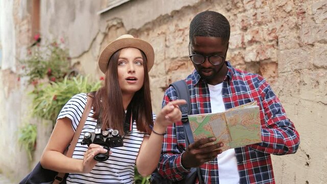 Young multinational couple of tourists standing on street and looking for necessary attractions on map taking photos on camera.