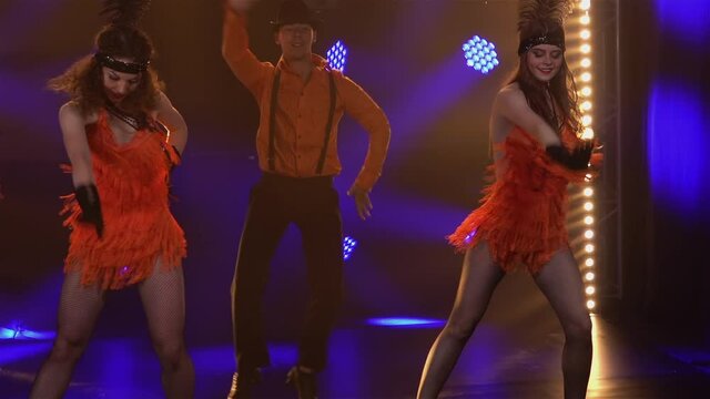 Dance group dressed in Chicago style costumes from 30s dancing cheerfully in studio with bright lights. Man in hat and woman in short dresses perform theatrical show at party. Close up. Slow motion.