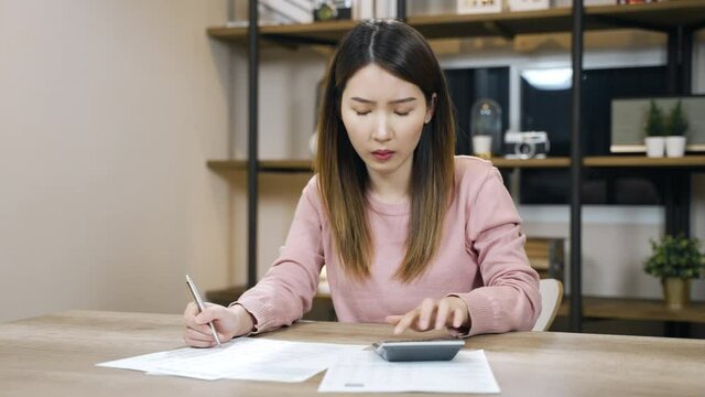Stressed Asian woman looking at the paper bills calculating credit card invoice or unpaid taxes and bank account overpay at home