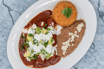 Overhead view of chicken enchiladas smothered in red sauce served with refried beans and rice for a tasty Mexican food meal.