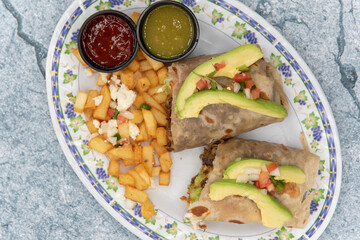 Overhead view of avacado slices top both halves of the flavorful Mexican breakfast burrito served with cubed potatoes and dipping sauce.