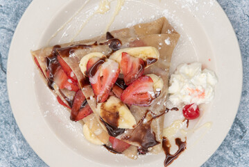 Overhead view of mouthwatering plated of crepes piled with sliced bananas and strawberrries topped with whipped cream and syrup.