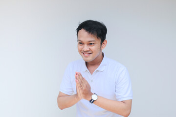 Young Asian man wearing white shirt give namaste greeting hands. Indonesian man on grey background.