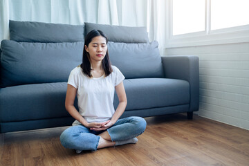 Beautiful Asian woman waking up in the morning practicing meditating exercise at home living room, routine healthy calm mind and body lifestyle spiritual mantra focus meditation lotus sitting position